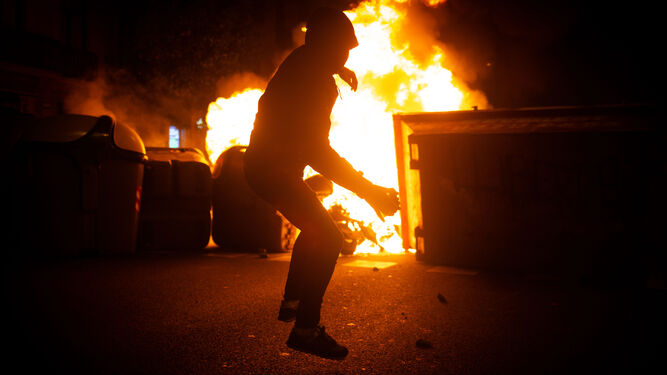 Un hombre se dispone a lanzar un objeto ante unos contenedores ardiendo