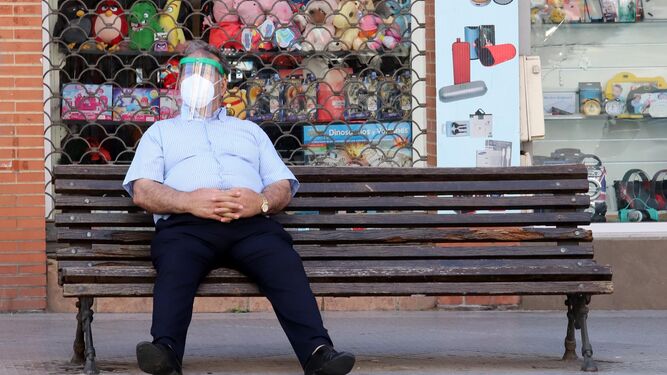 Un hombre se sienta en un banco con una mascarilla y una pantalla de protección.